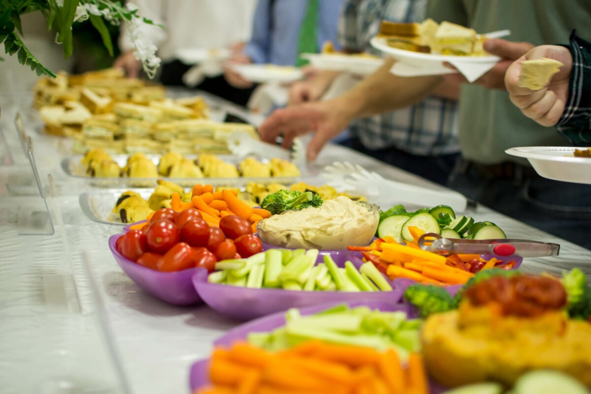 Photo shows a buffet of colorful vegetables, salad, and other foods as people queue to load up their plates