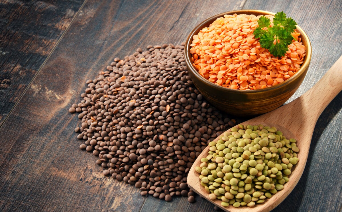 Photo shows three different varieties of lentil on a wooden surface: brown, on the table; red, in a small bowl; and green, in a wooden spoon