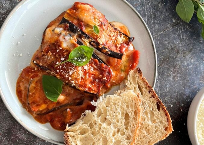 A plate of vegan and dairy-free aubergine parmigiana served alongside white bread and vegan parmesan