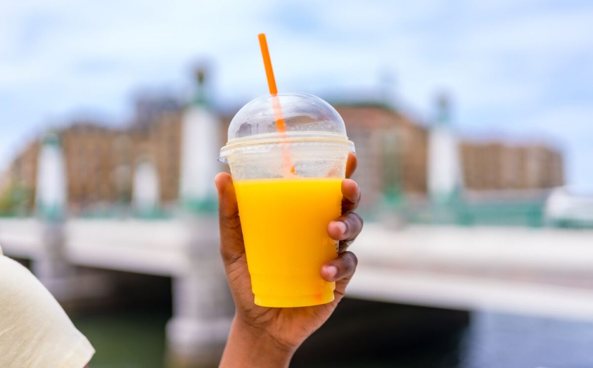 Photo shows someone holding up a plastic cup with a straw full of orange juice