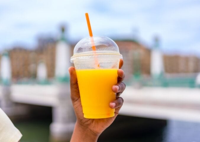 Photo shows someone holding up a plastic cup with a straw full of orange juice