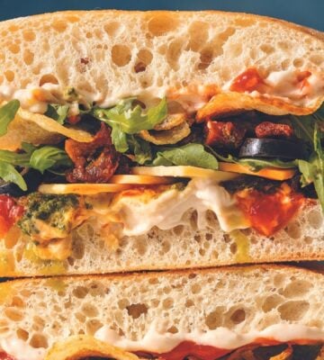 a vegan sandwich filled with pesto, hummus, potato chips, jalepenos, olives, arugula, vegan mayo, and cheese on a ciabatta loaf