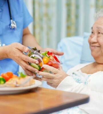 Photo shows a healthcare worker handing an older patient a bowl of brightly colored salad while she reclines in a hospital bed