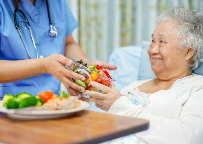 Photo shows a healthcare worker handing an older patient a bowl of brightly colored salad while she reclines in a hospital bed