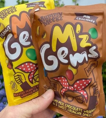 Two packets of "M'z Gems" (vegan dupes of M&Ms) being held up in front of a flower bush