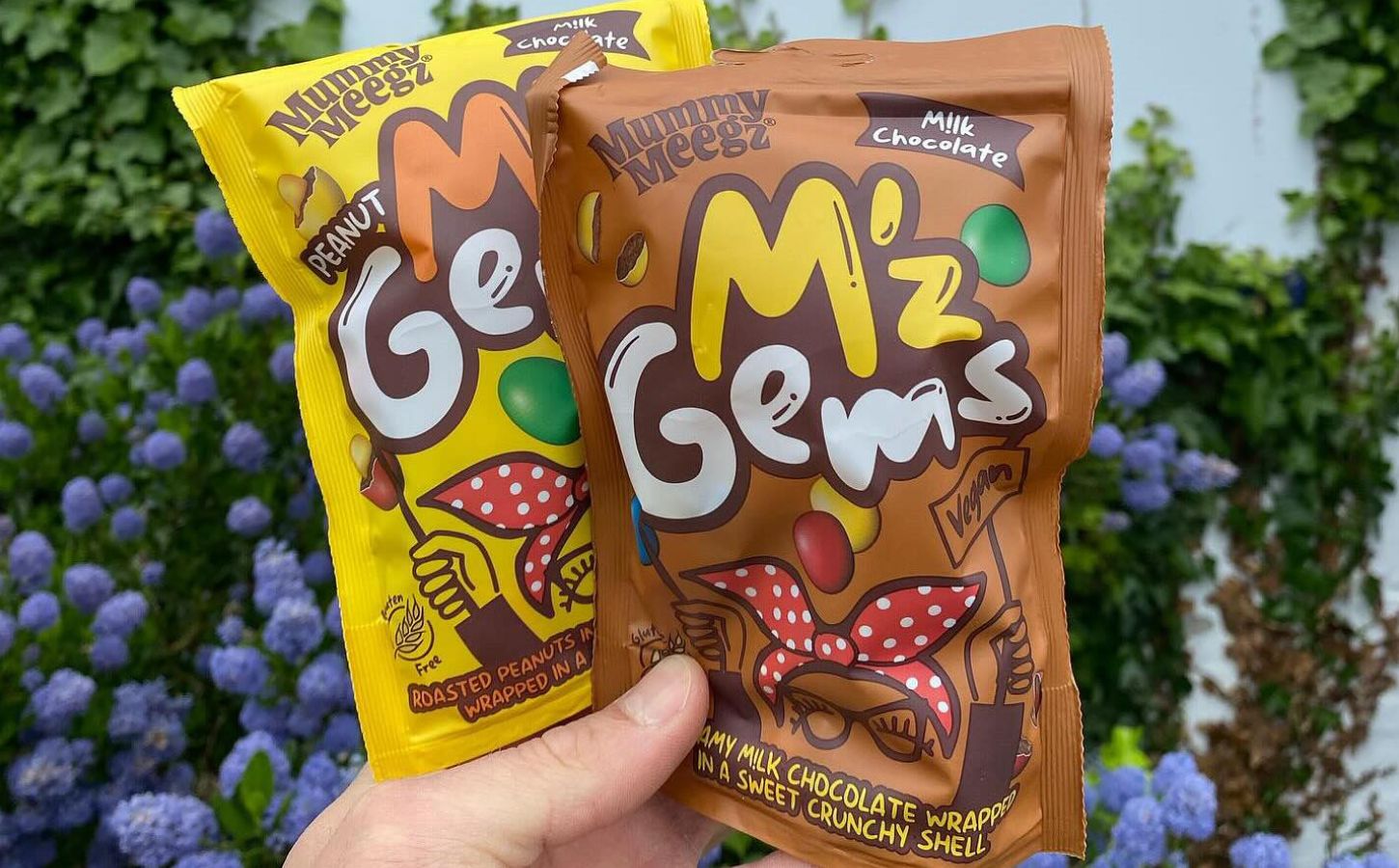 Two packets of "M'z Gems" (vegan dupes of M&Ms) being held up in front of a flower bush