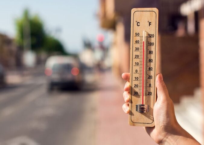 Photo shows someone's hand as they hold up a wooden thermometer on a hot city street - the mercury shows over 40C