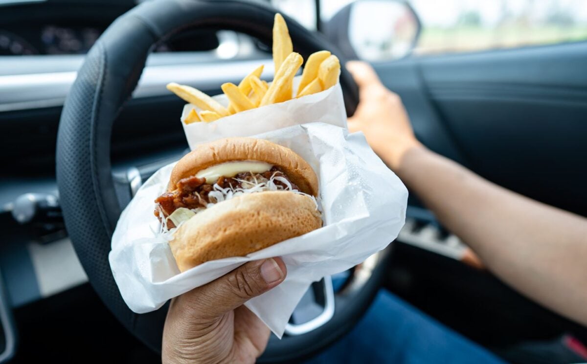 Photo shows someone driving a car and eating a hamburger and fries at the same time.