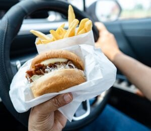 Photo shows someone driving a car and eating a hamburger and fries at the same time.
