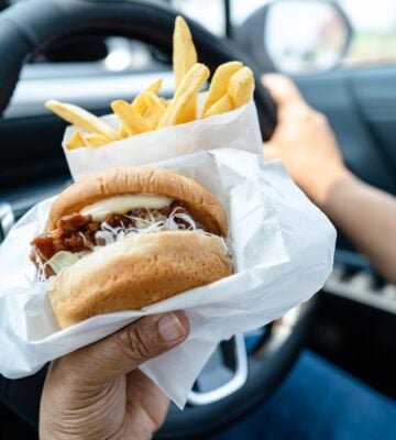 Photo shows someone driving a car and eating a hamburger and fries at the same time.