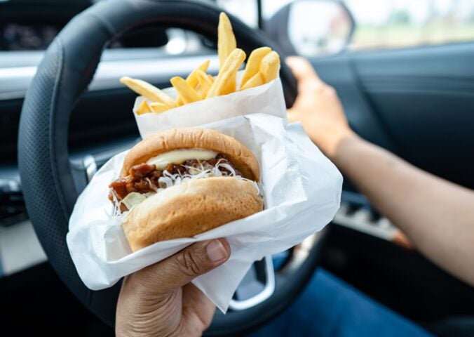 Photo shows someone driving a car and eating a hamburger and fries at the same time.