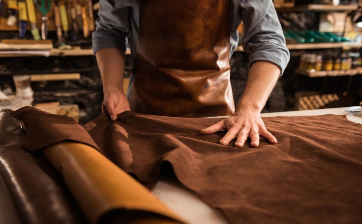 Photo shows a cobbler in a leather apron working with large sheets of hide in a shop