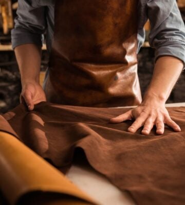 Photo shows a cobbler in a leather apron working with large sheets of hide in a shop