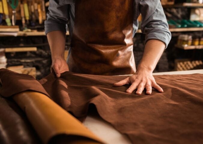 Photo shows a cobbler in a leather apron working with large sheets of hide in a shop