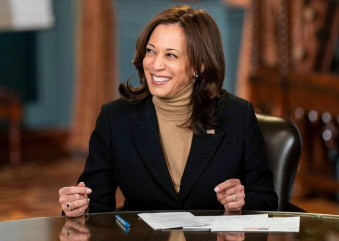 Photo shows Kamala Harris seated at a wooden table and smiling off-camera