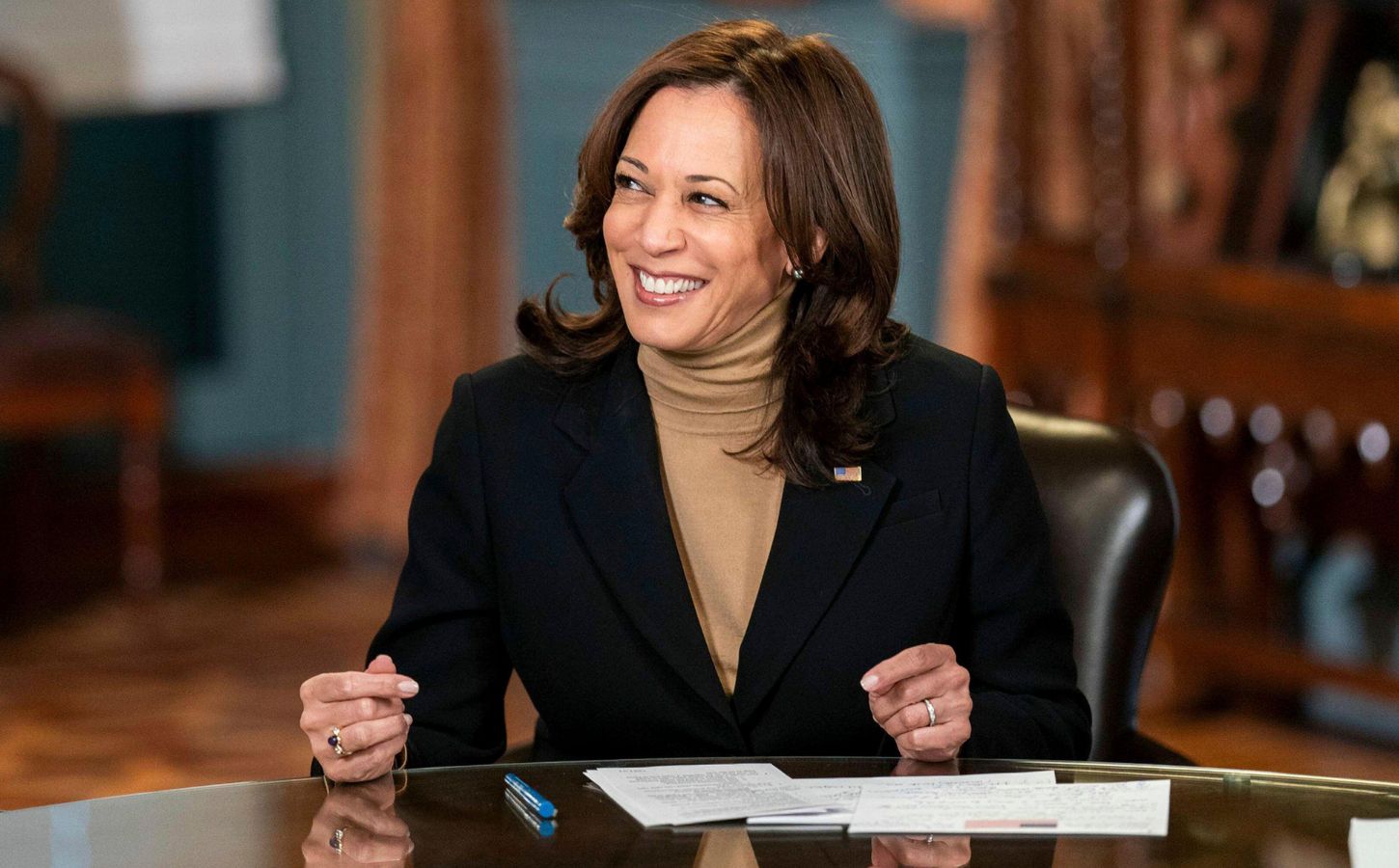 Photo shows Kamala Harris seated at a wooden table and smiling off-camera