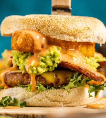 a picture of a stacked vegan breakfast burger made with vegan burger patties, vegan smoky bacon, vegan cheddar, guacamole, and more topped on an English muffin