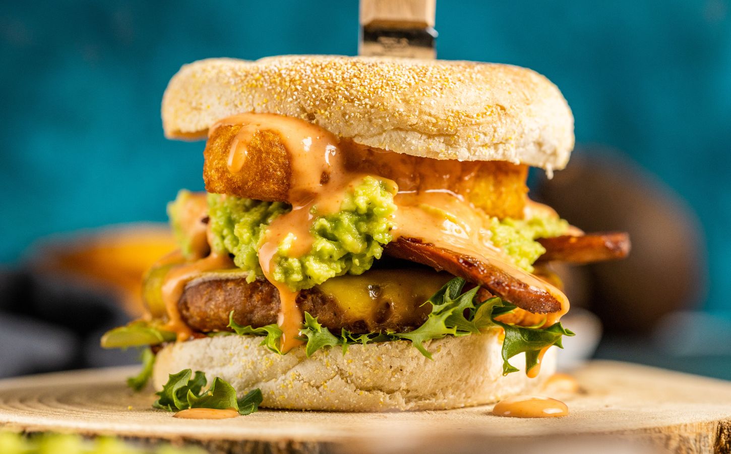 a picture of a stacked vegan breakfast burger made with vegan burger patties, vegan smoky bacon, vegan cheddar, guacamole, and more topped on an English muffin