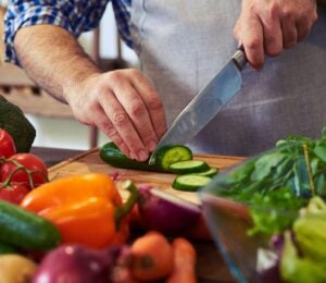 Photo shows someone standing in a kitchen, wearing an apron, and chopping fresh fruit and vegetables