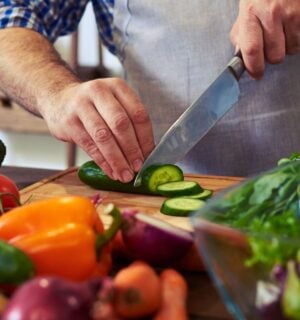 Photo shows someone standing in a kitchen, wearing an apron, and chopping fresh fruit and vegetables