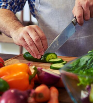 Photo shows someone standing in a kitchen, wearing an apron, and chopping fresh fruit and vegetables