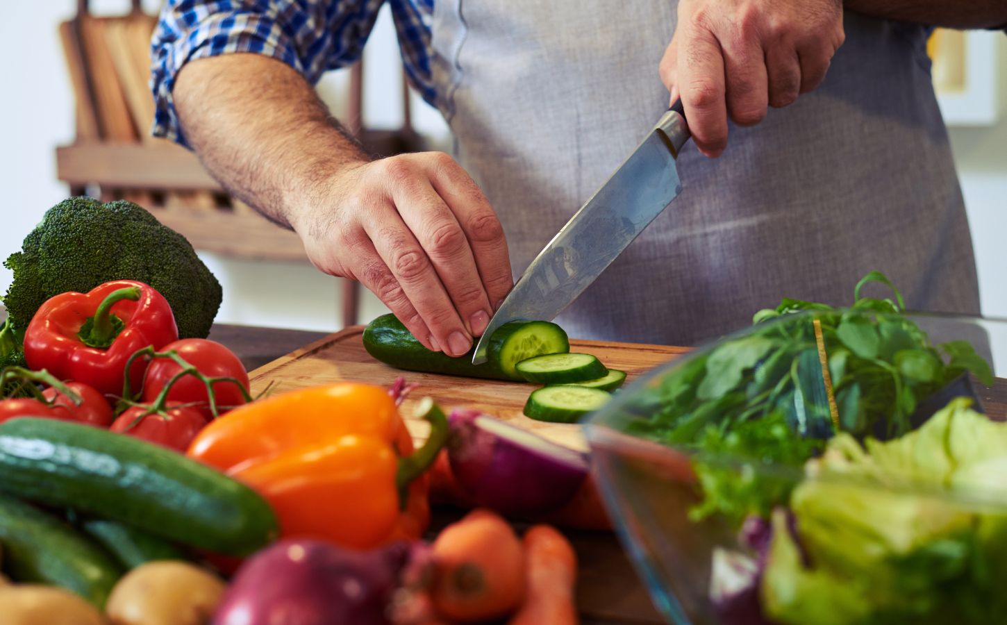 Photo shows someone standing in a kitchen, wearing an apron, and chopping fresh fruit and vegetables