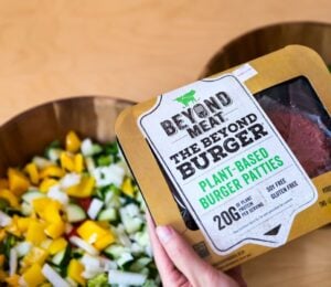 Photo shows someone's hands as they hold up a pack of Beyond Meat burger patties above two bowls of chopped vegetables