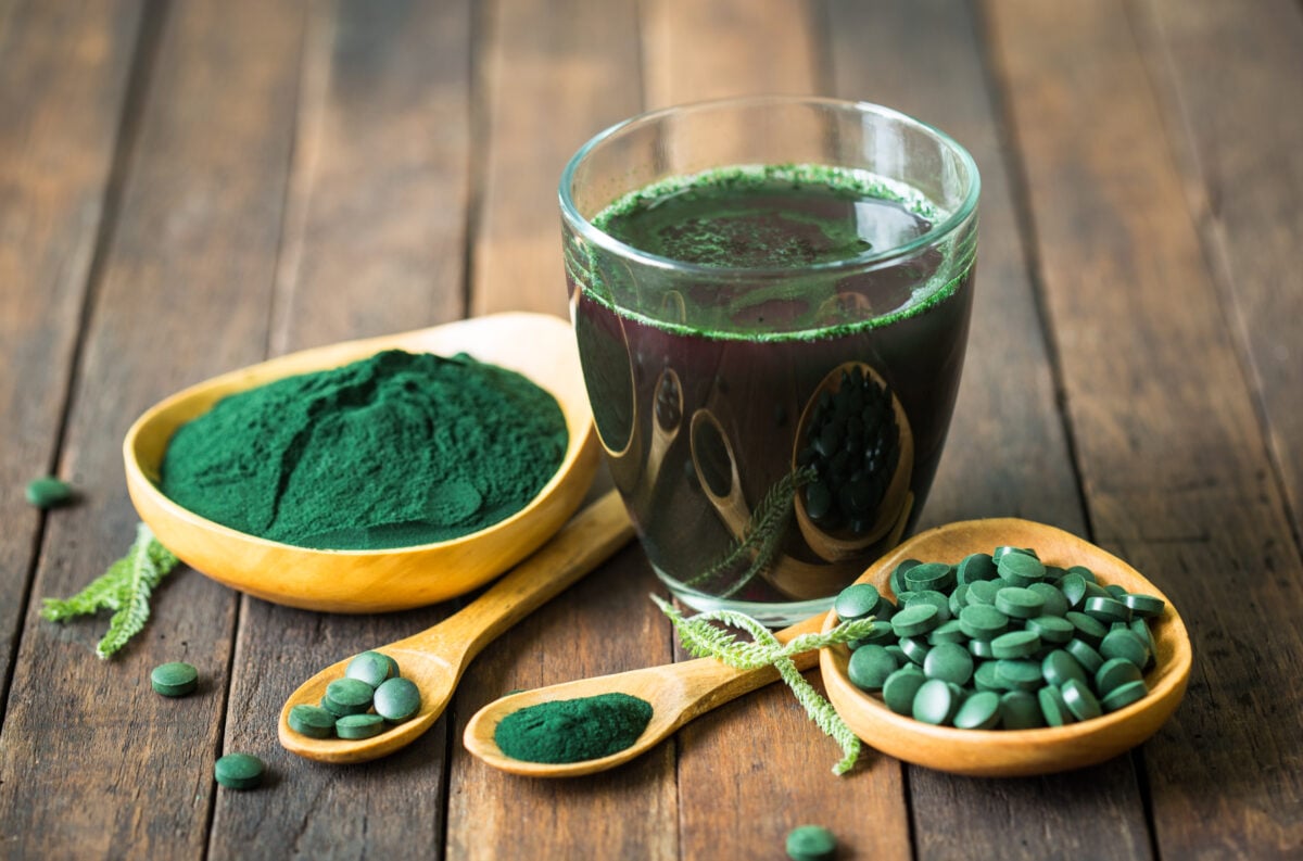 A green glass of nutrient dense spirulina, which carries a number of health benefits, next to some spirulina powder and pills