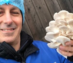 Photo shows chef Derek Sarno wearing PPE and holding up a handful of bright white, layered mushrooms to the camera