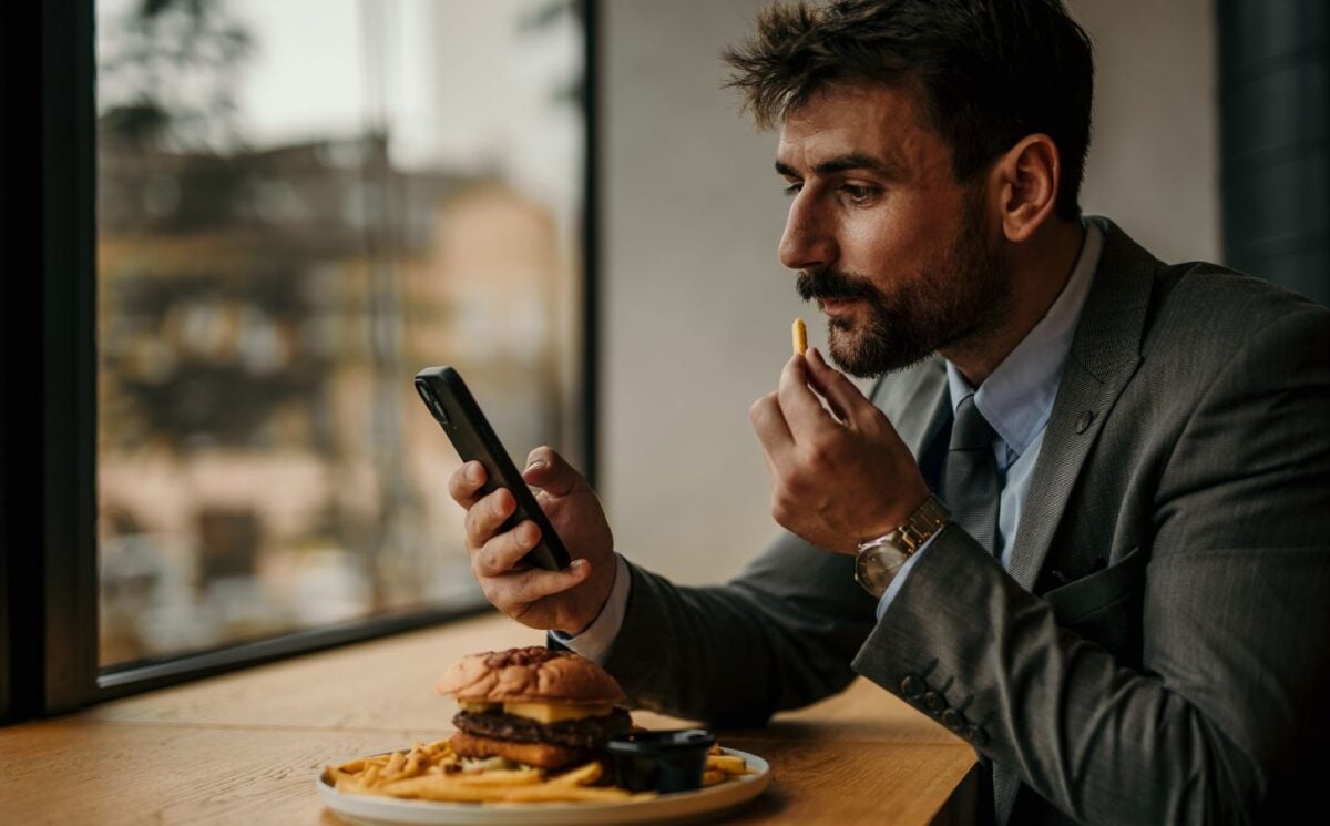 Photo shows a man sitting at a table and eating while he uses his smartphone