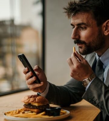 Photo shows a man sitting at a table and eating while he uses his smartphone