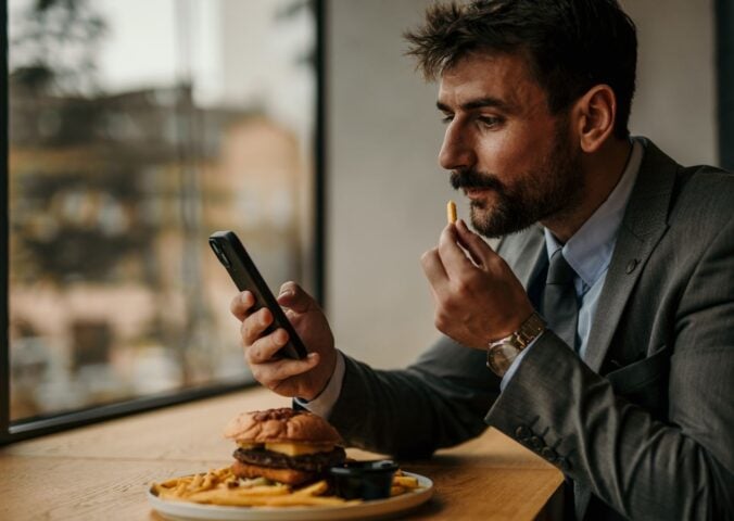 Photo shows a man sitting at a table and eating while he uses his smartphone