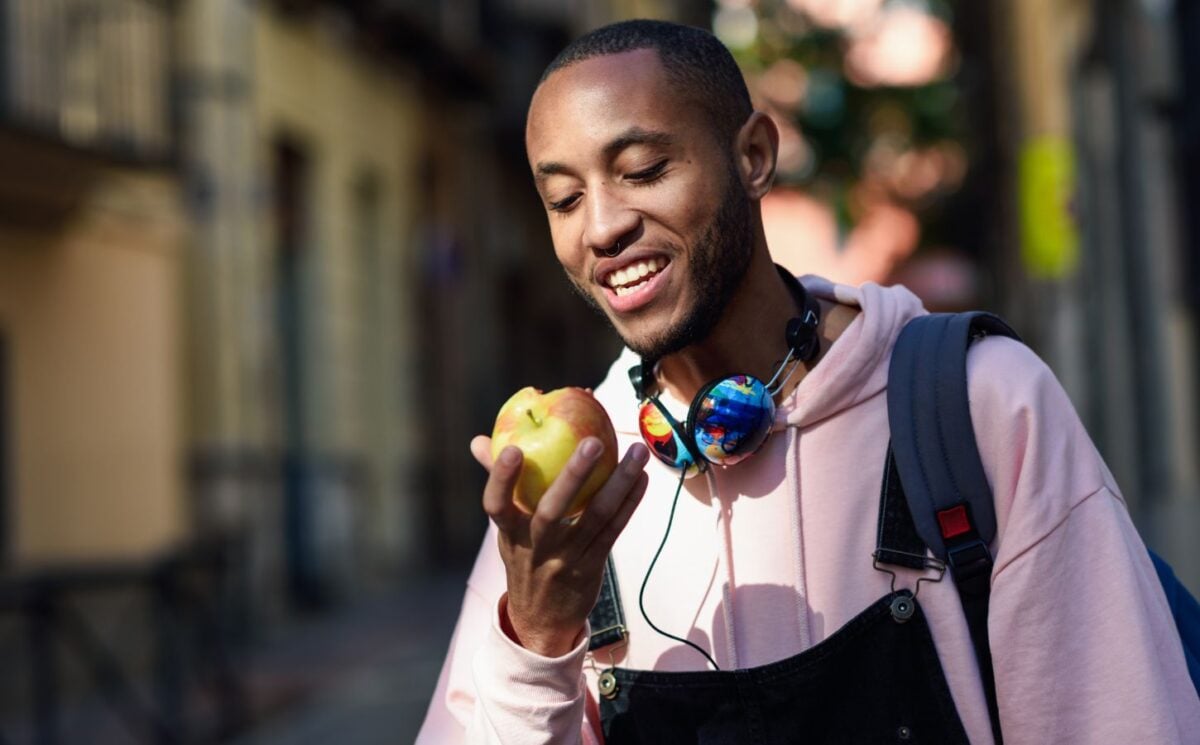 A vegan man in a pink hoody and dungarees eating an apple