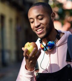 A vegan man in a pink hoody and dungarees eating an apple