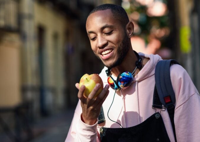 A vegan man in a pink hoody and dungarees eating an apple