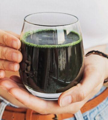 A woman holding a glass of dark green liquid containing cultivated spirulina, which is a good vegan source of B12