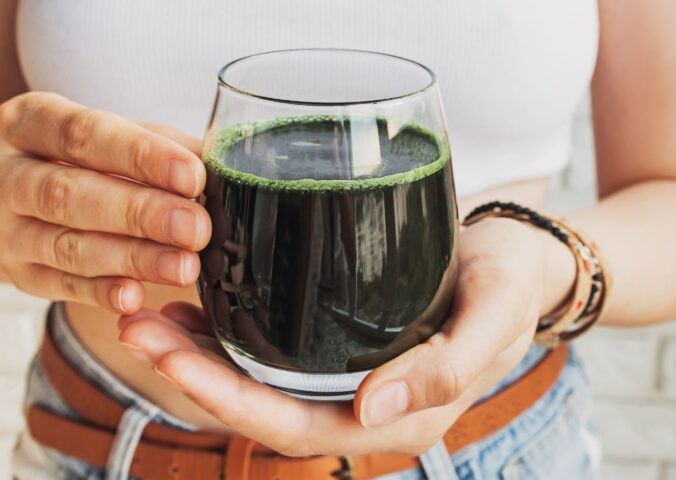 A woman holding a glass of dark green liquid containing cultivated spirulina, which is a good vegan source of B12