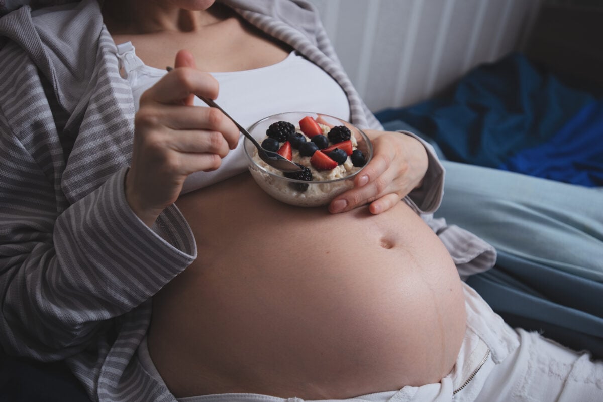 A close up of a pregnant woman's tummy as she eats porridge with fruit in bed