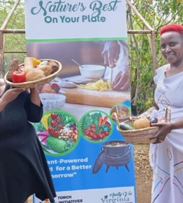 Photo shows Virginia Ruguru (left) and Camp Gedeng manager Bibi Kui (right) holding up bowls of produce in front of a large sign promoting plant-based foods and Africa Vegan Restaurant Week