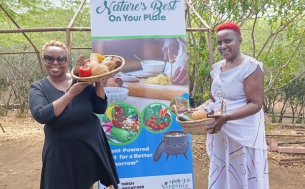 Photo shows Virginia Ruguru (left) and Camp Gedeng manager Bibi Kui (right) holding up bowls of produce in front of a large sign promoting plant-based foods and Africa Vegan Restaurant Week