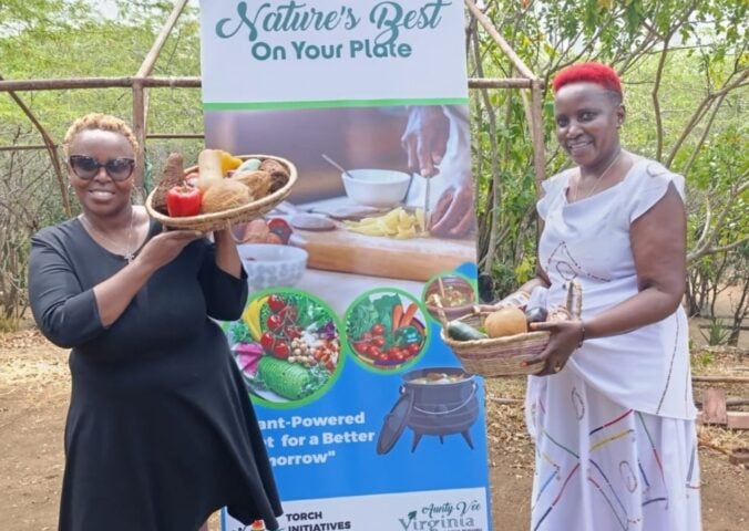 Photo shows Virginia Ruguru (left) and Camp Gedeng manager Bibi Kui (right) holding up bowls of produce in front of a large sign promoting plant-based foods and Africa Vegan Restaurant Week