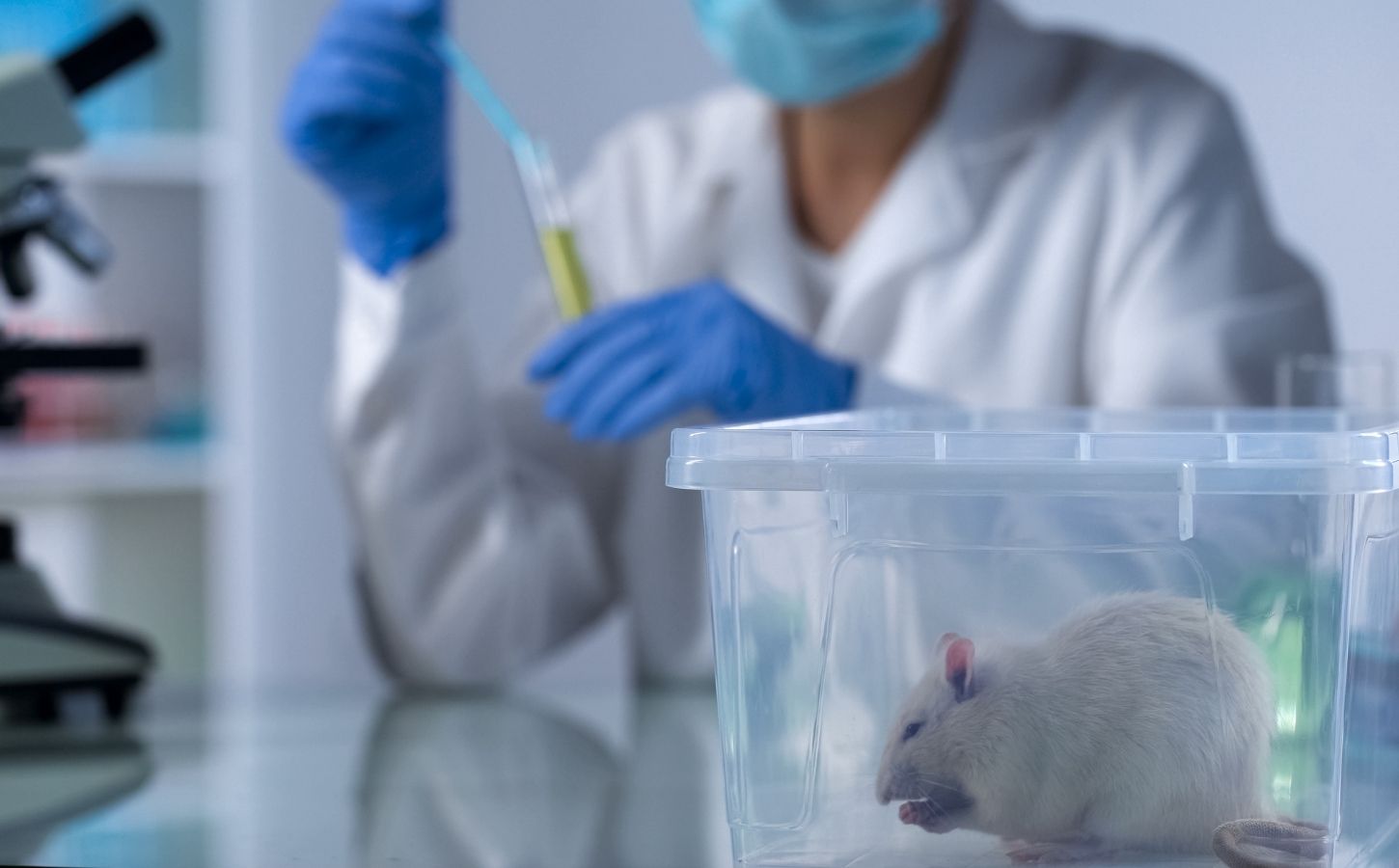 Photo shows a scientist in a white coat and blue globes filling a syring in the background while a white mouse sits in a plastic box in the foreground