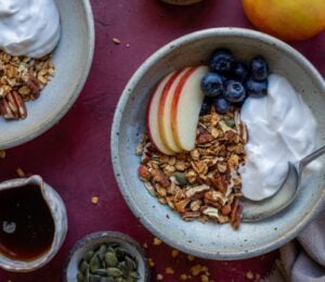 A bowl of vegan apple granola alongside apple slices, blueberry, and dairy-free yogurt