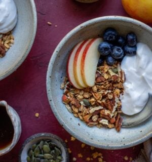 A bowl of vegan apple granola alongside apple slices, blueberry, and dairy-free yogurt