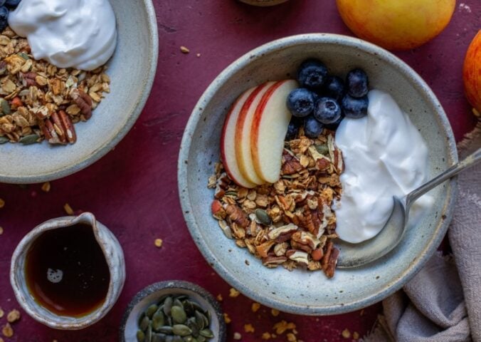 A bowl of vegan apple granola alongside apple slices, blueberry, and dairy-free yogurt