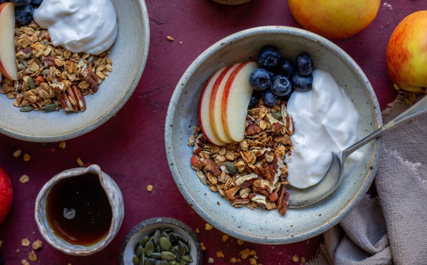 A bowl of vegan apple granola alongside apple slices, blueberry, and dairy-free yogurt