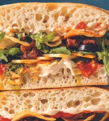 a picture of an everything sarnie filled with pesto, hummus, potato chips, jalapenos, olives, arugula, vegan mayo, and cheese on a ciabatta loaf