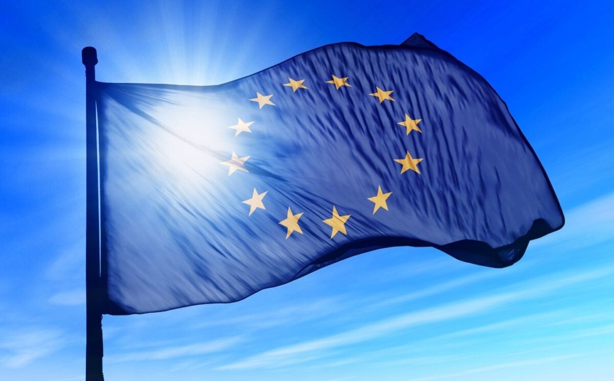 The European flag in front of a bright sun and blue sky, with the sun shining through it