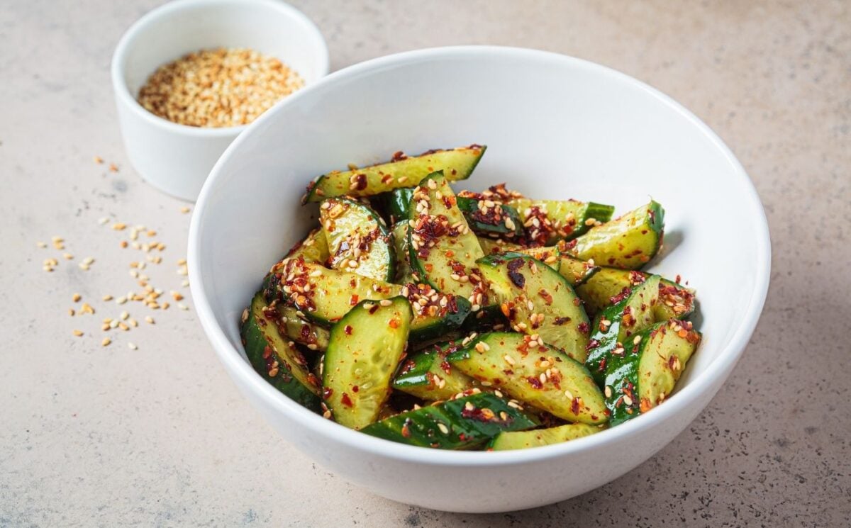 A white bowl of vegan cucumber salad covered in chili next to a white pot of sesame seeds