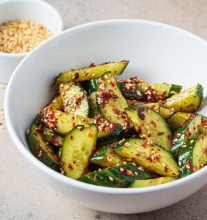 A white bowl of vegan cucumber salad covered in chili next to a white pot of sesame seeds